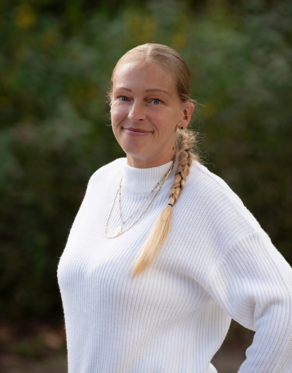 Frau mit langen blonden Zopf mit weißem Pullover steht vor einem schwarzen Hintergrund.