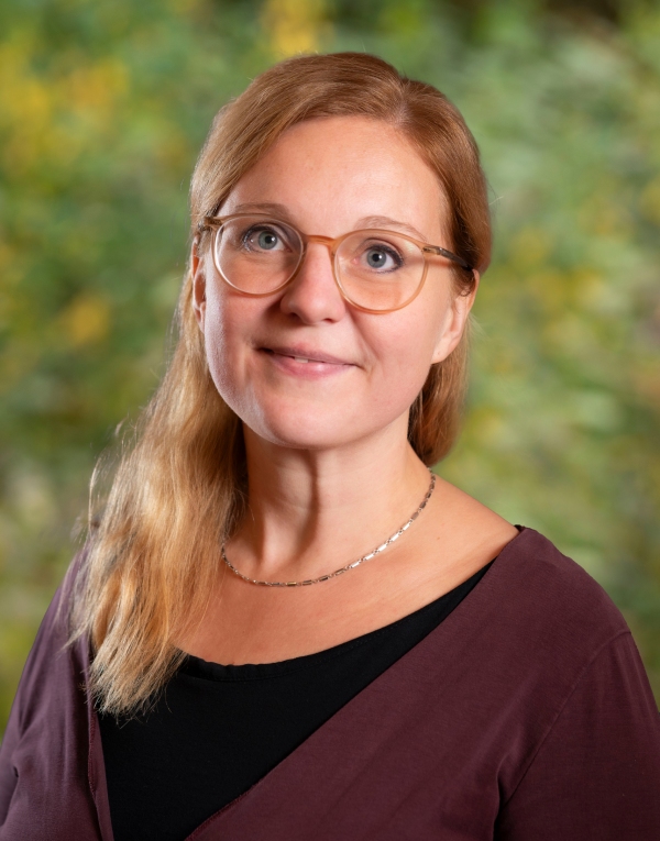 Frau mit schulterlangem Haar und Brille trägt einen schwarzen Pullover und steht vor einem grünen Hintergrund.