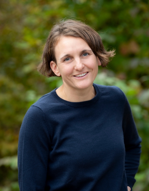 Frau mit braunen kurzen Haaren und einem Blauen Pullover steht vor einem grünen Hintergrund und lächelt.