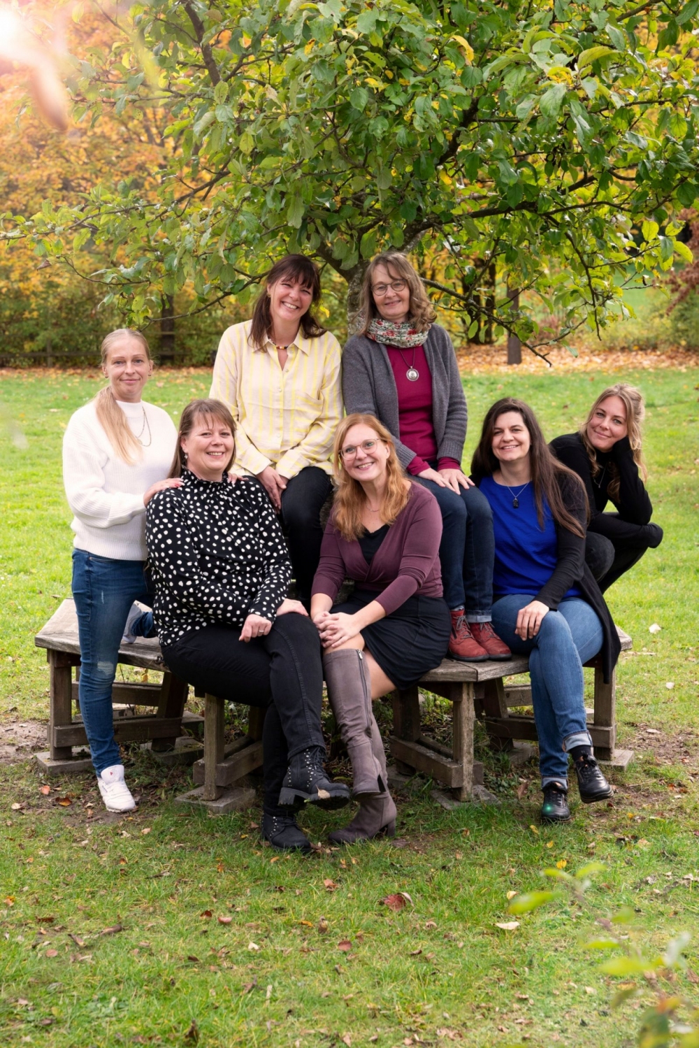 Sieben Frauen sitzen und stehen auf einer hölzernen Sitzgruppe im Freien. Im Hintergrund ist ein Baum mit grünen Blättern zu sehen, das Gras ist frisch und die Umgebung wirkt herbstlich. Die Frauen tragen unterschiedliche Kleidung, darunter Pullover, Blusen und Jeans. Einige lächeln, andere schauen neutral in die Kamera. Die Szene wirkt freundlich und entspannt.