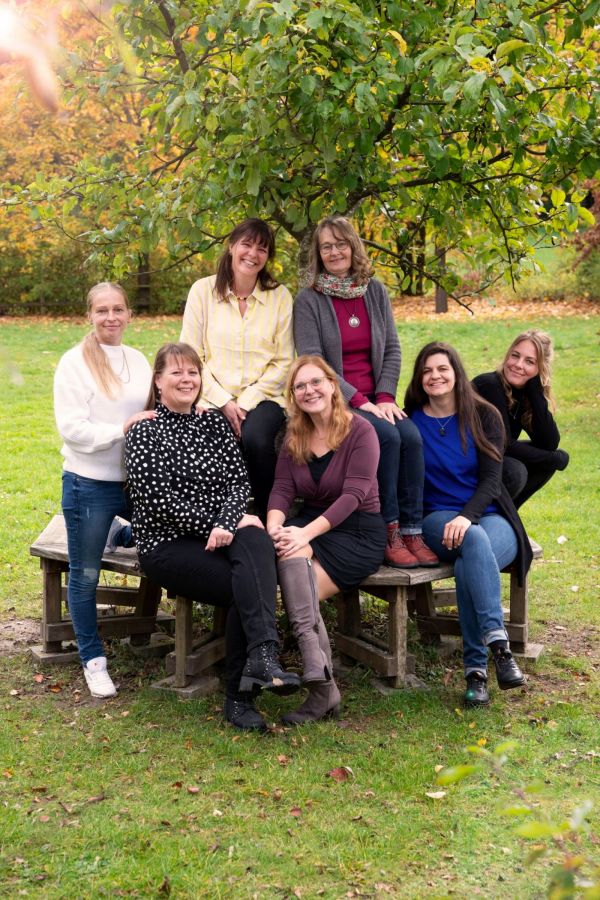 Sieben Frauen sitzen und stehen auf einer hölzernen Sitzgruppe im Freien. Im Hintergrund ist ein Baum mit grünen Blättern zu sehen, das Gras ist frisch und die Umgebung wirkt herbstlich. Die Frauen tragen unterschiedliche Kleidung, darunter Pullover, Blusen und Jeans. Einige lächeln, andere schauen neutral in die Kamera. Die Szene wirkt freundlich und entspannt.