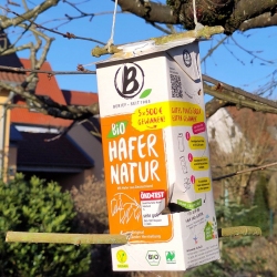 Ein rechteckiger Karton mit orange-grüner Aufschrift hängt mithilfe von zwei Schnüren an einem Ast. Der Karton dient als improvisiertes Futterhäuschen und ist mit zwei quer durchgesteckten Holzstäben versehen, die als Sitzstangen für Vögel dienen. Im Hintergrund sind ein Haus mit Dach und ein Garten zu sehen.